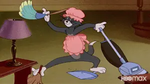 Tom the cat, wearing a pink apron and hat, frantically cleans with a broom, dustpan, and vacuum in a living room.