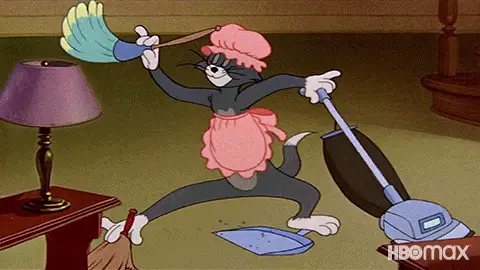 Tom the cat, wearing a pink apron and hat, frantically cleans with a broom, dustpan, and vacuum in a living room.
