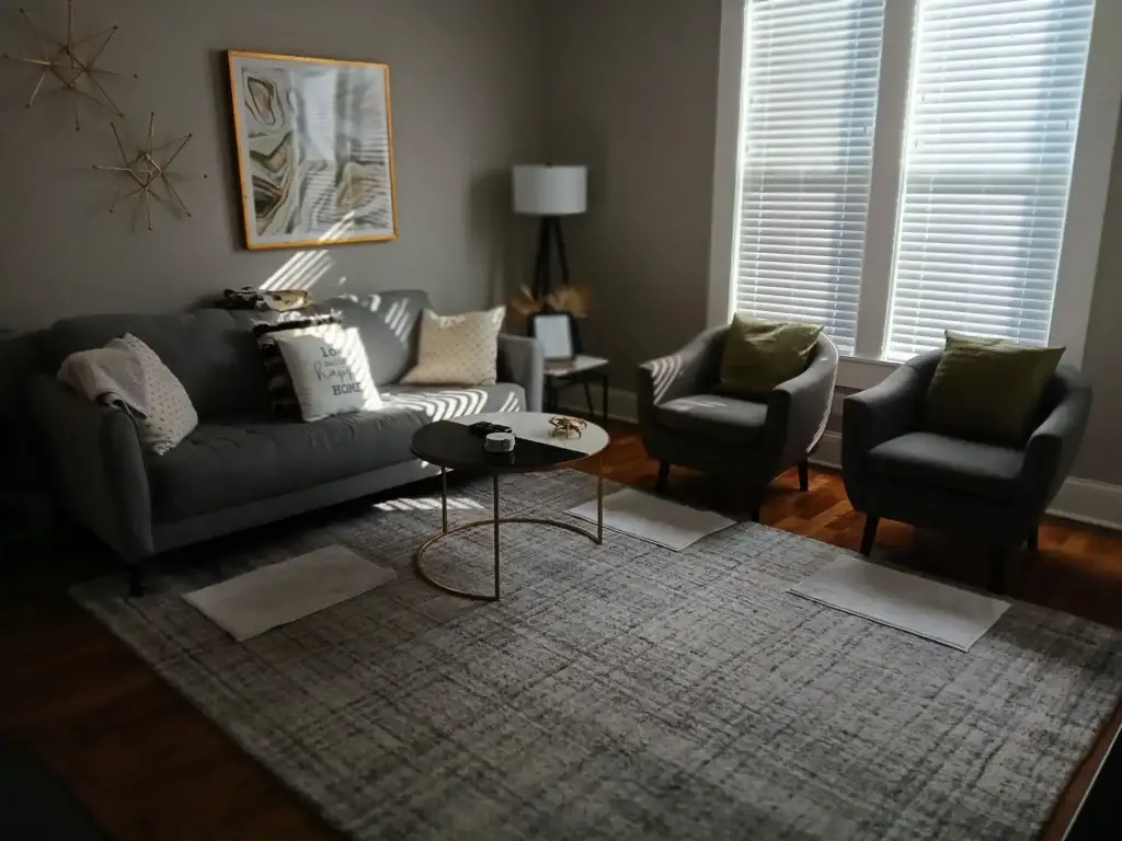 A modern living room with a gray sofa, two gray chairs, a coffee table, and sunlight streaming through window blinds.