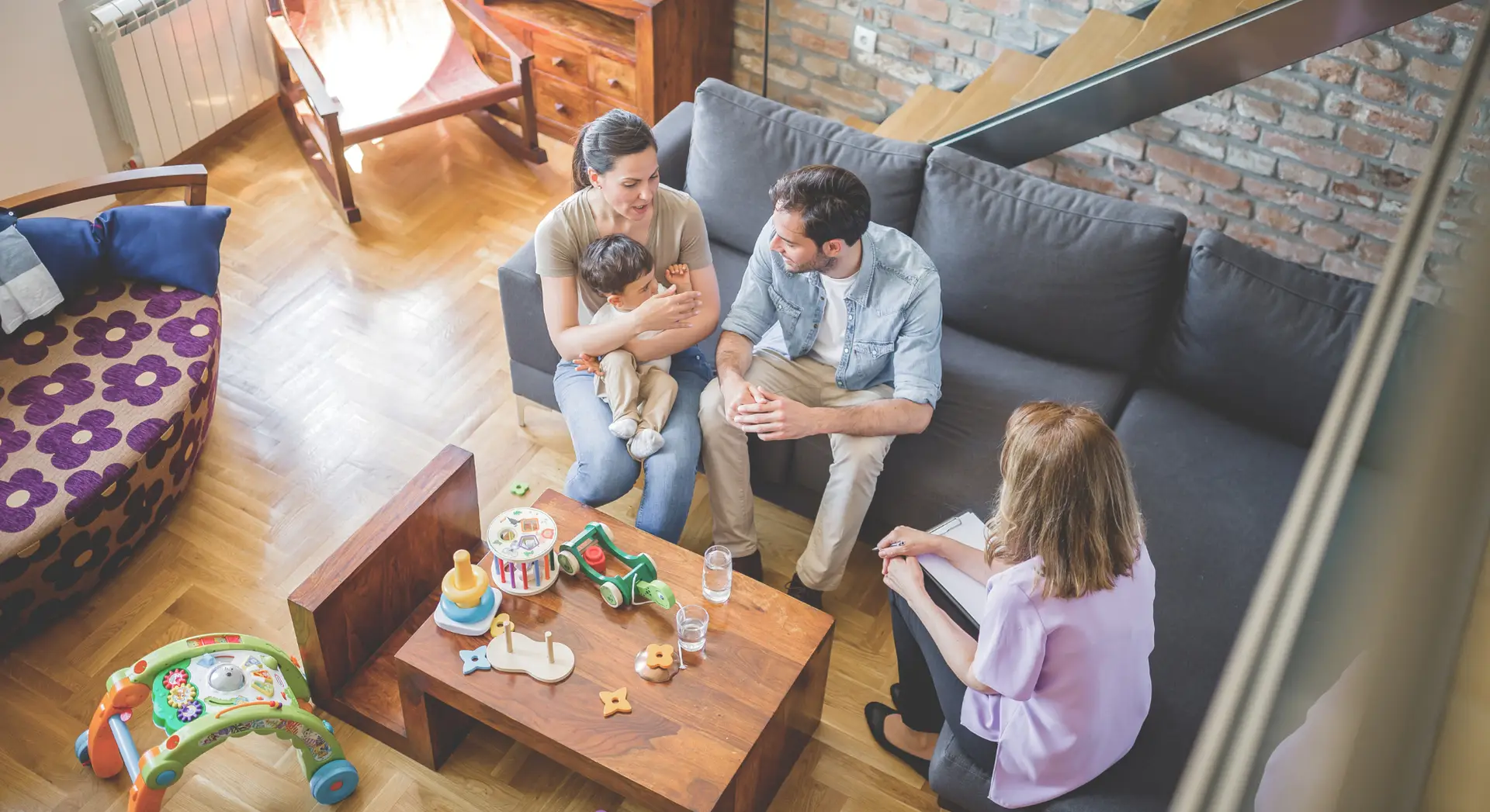 A family with a baby talks to a woman with a notepad while sitting in a living room with toys and drinks on the table.