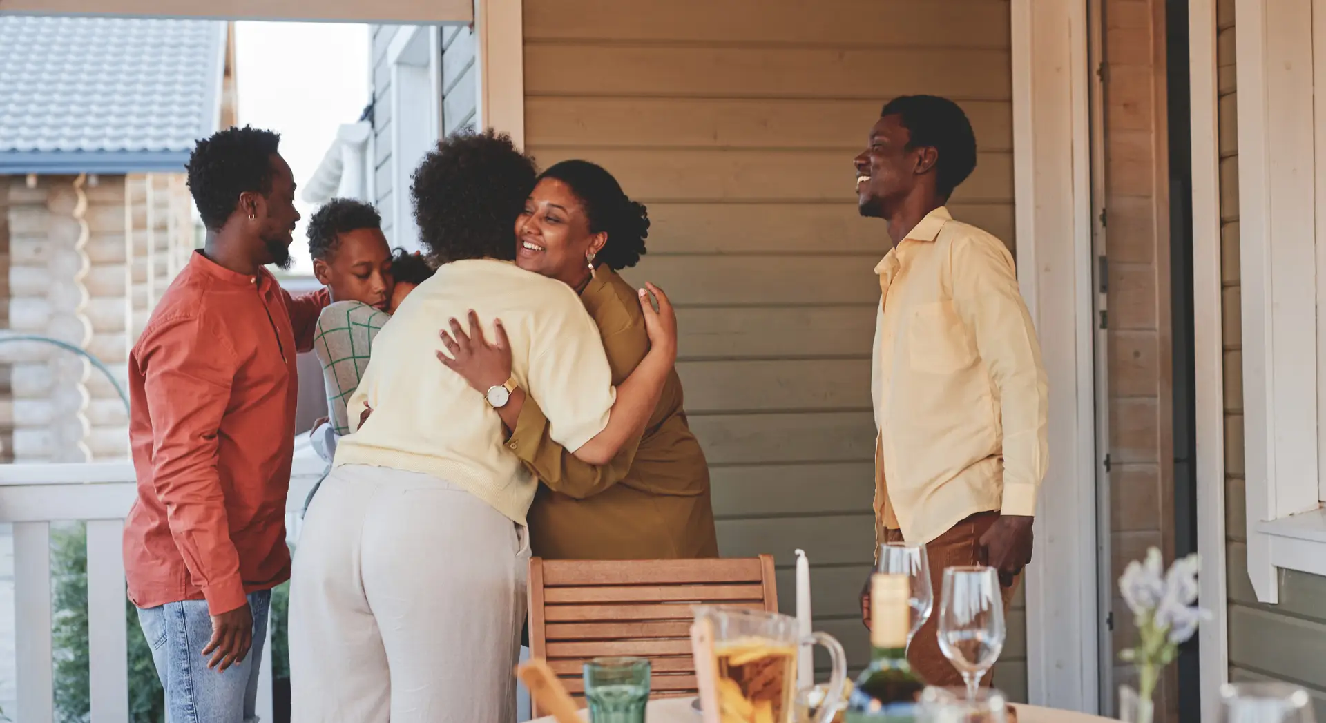 A group of people greet each other with hugs and smiles on a porch, gathered around a table set for a meal.