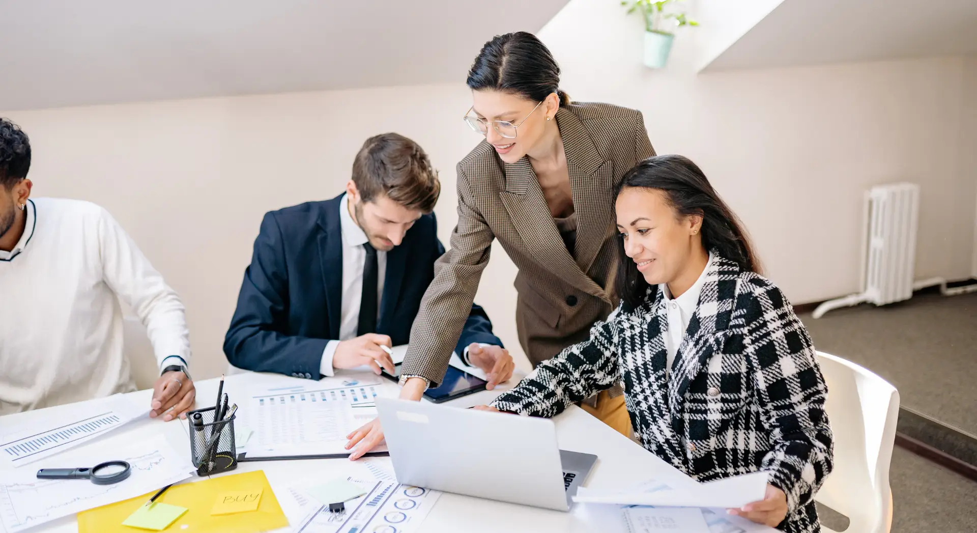 Three people in business attire working together at a desk with papers, charts, and a laptop in a bright office.