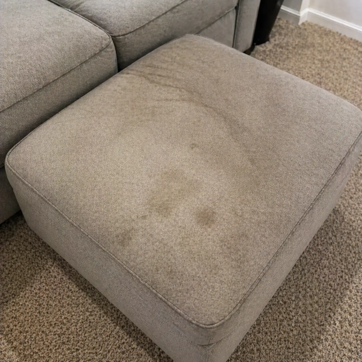 A gray upholstered ottoman with visible water stains sits on a beige carpet next to a matching sofa.