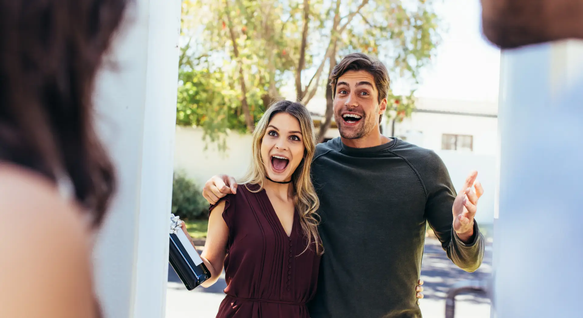 A smiling couple stands at a doorway, looking surprised and happy to see the people inside.