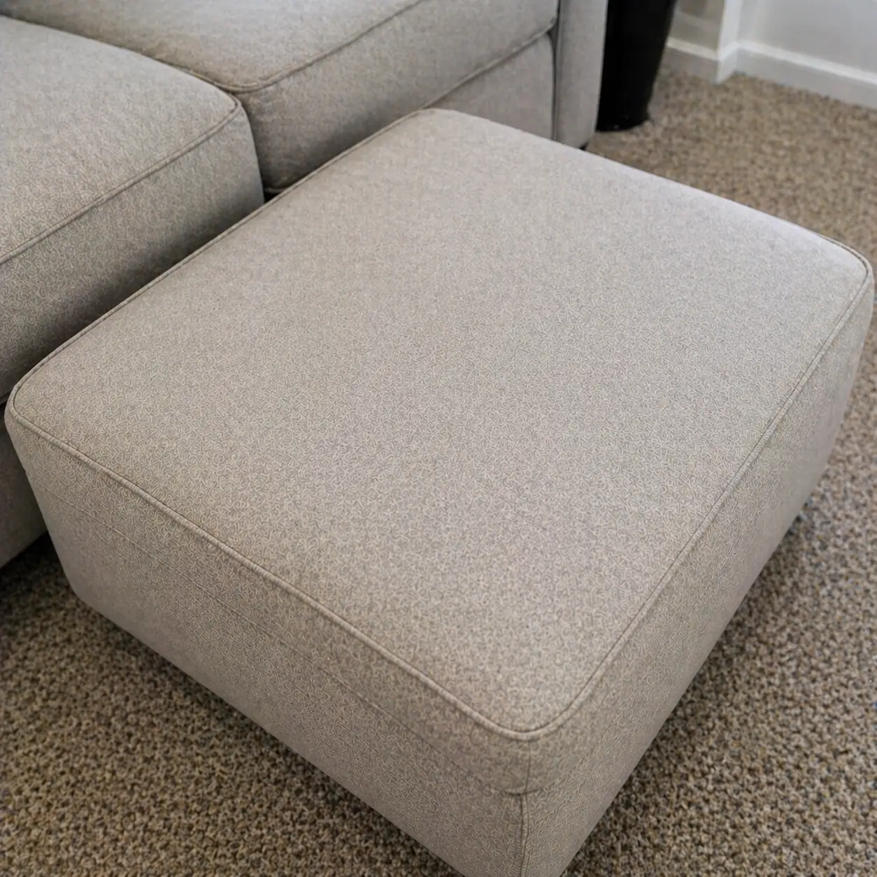 A light gray upholstered ottoman on a speckled carpet next to a matching sofa.