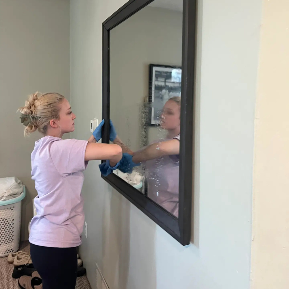 A woman in a purple shirt cleans a large wall mirror with a blue cloth in a home setting.