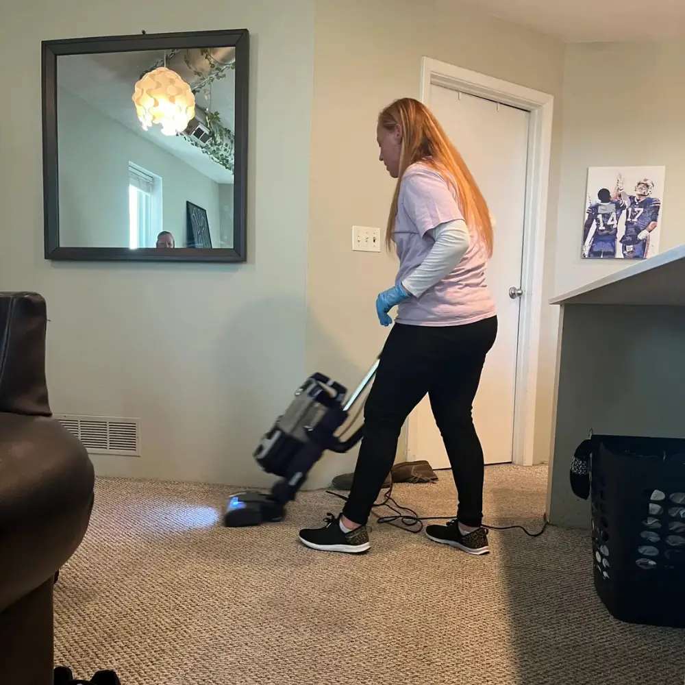 Woman vacuuming a carpeted living room, with a laundry basket and wall art visible in the background.
