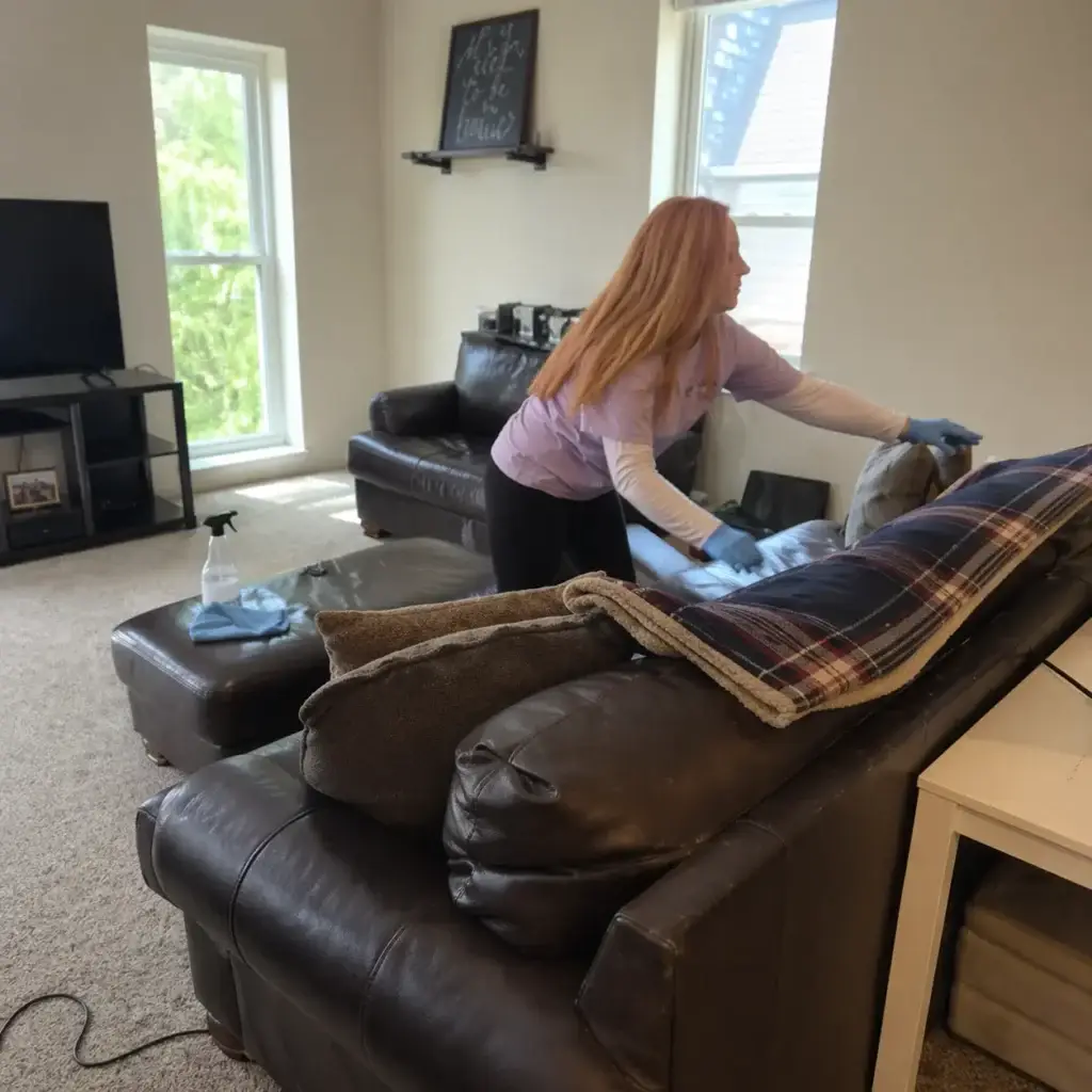 A woman wearing gloves cleans a leather couch in a bright living room with large windows and a TV.