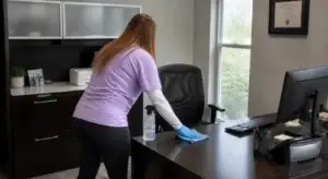 A woman wearing gloves cleans an office desk with a cloth and spray bottle near a computer and window.