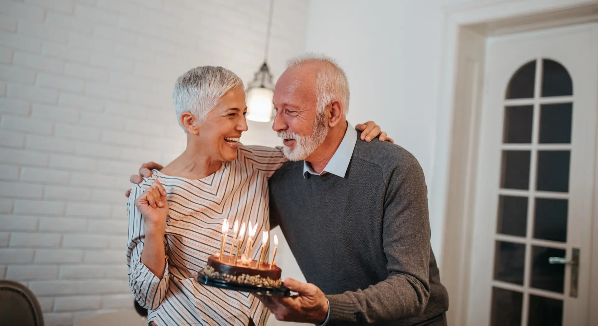Smiling older couple celebrates with a birthday cake topped with lit candles in a cozy, well-lit room.