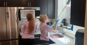 Two women cleaning a kitchen; one wipes the counter, while the other dusts near a window with a long duster.