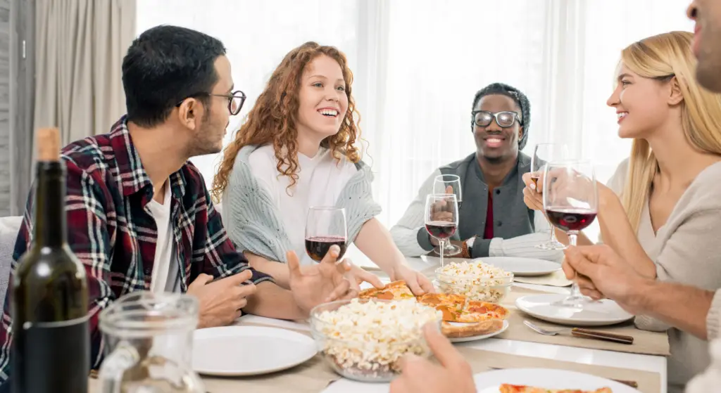 Five friends sit around a table, smiling and enjoying pizza, popcorn, and glasses of red wine together.