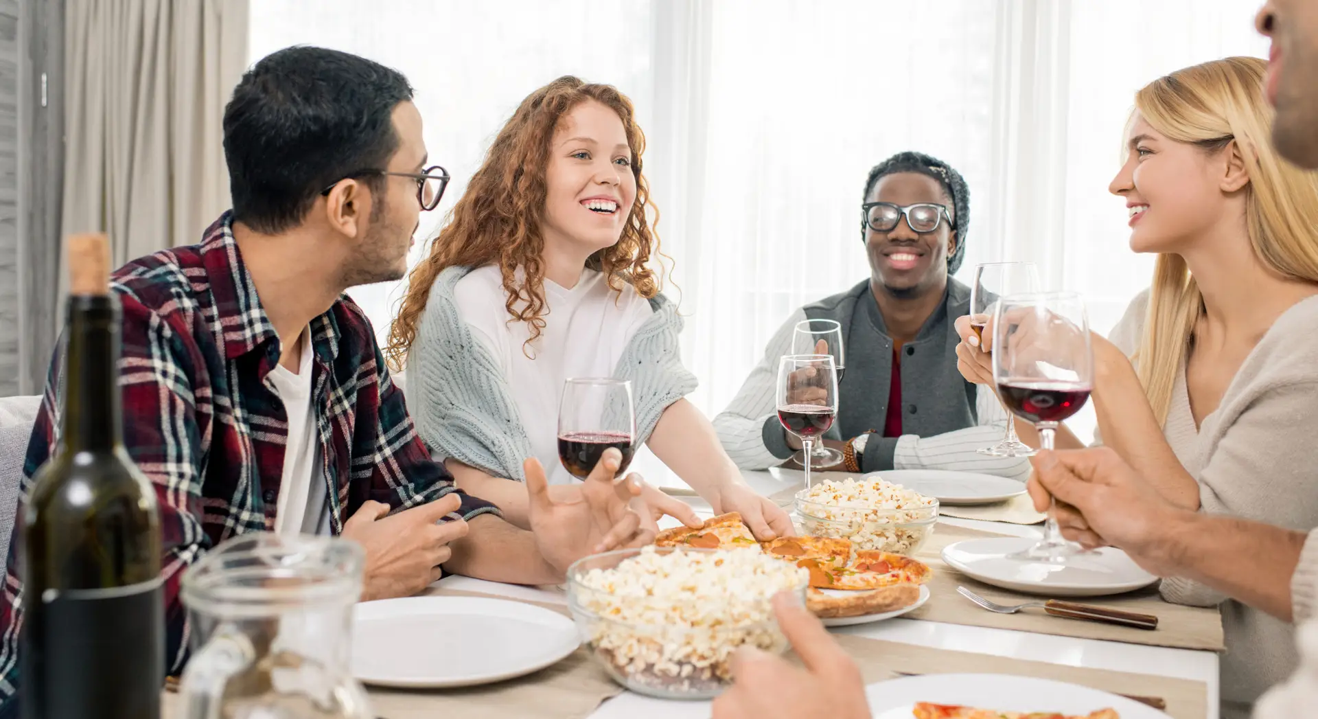 Five friends sit around a table, smiling and enjoying pizza, popcorn, and glasses of red wine together.