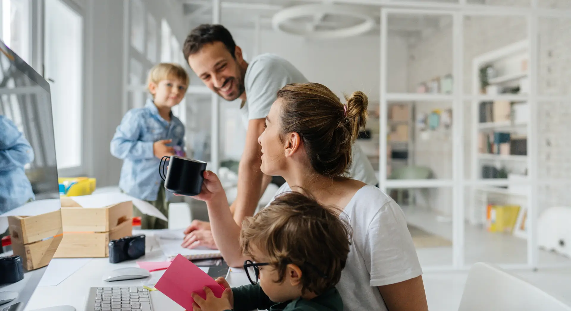 A family of four smiles and interacts together while working and playing at a desk in a bright home office.