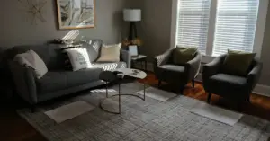 Modern living room with gray sofa, two armchairs, round coffee table, rug, and sunlight streaming through blinds.