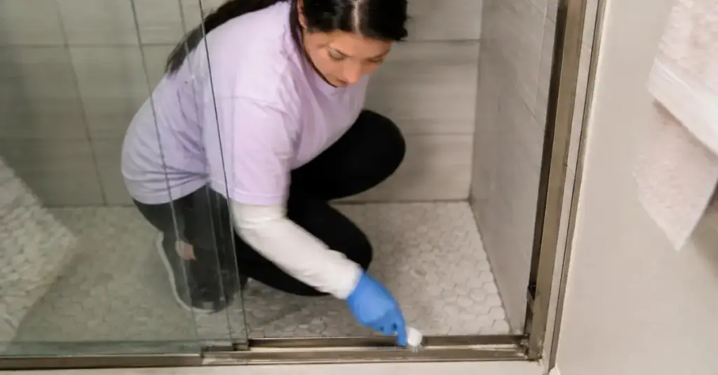 Person wearing a glove scrubbing the floor and edges of a shower with white tile.
