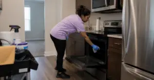 Woman in a purple shirt and blue gloves cleaning an open kitchen oven.