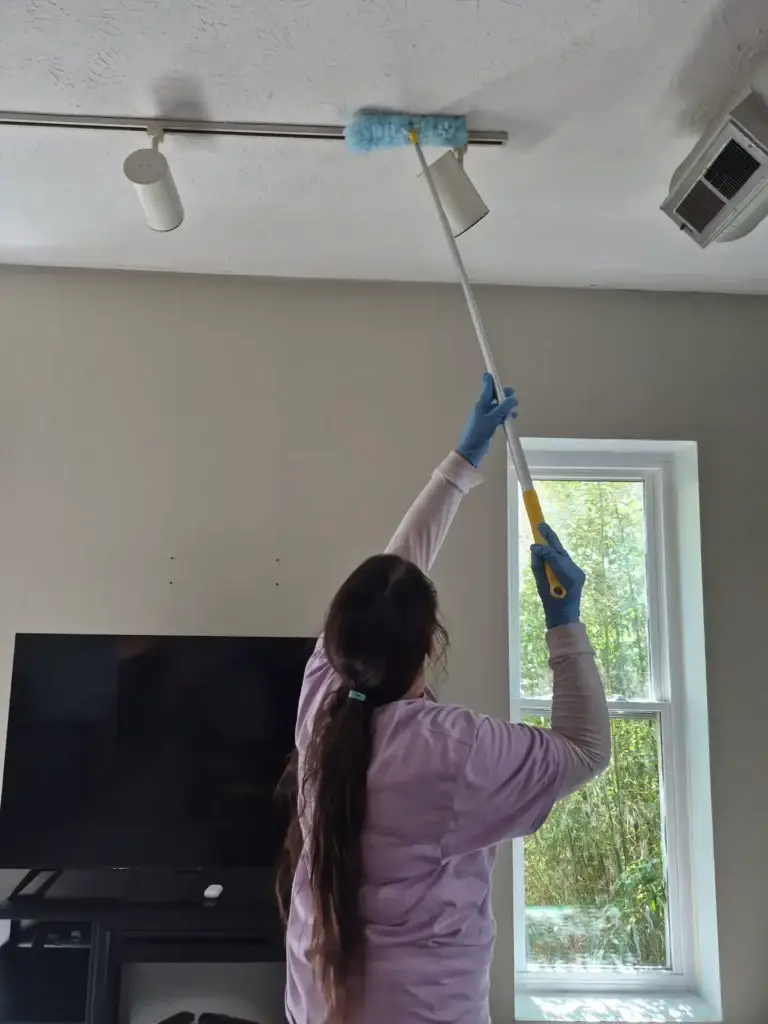 A person using a duster on a long pole to clean a ceiling light fixture in a living room.