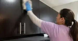 A woman in a purple shirt cleans dark kitchen cabinets with a cloth and blue gloves.