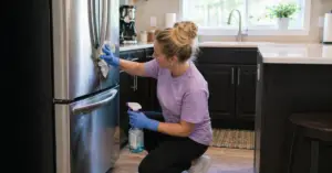 Woman kneeling by a stainless steel fridge, wiping the door with a cloth while holding a spray bottle wearing blue gloves in a bright kitchen.