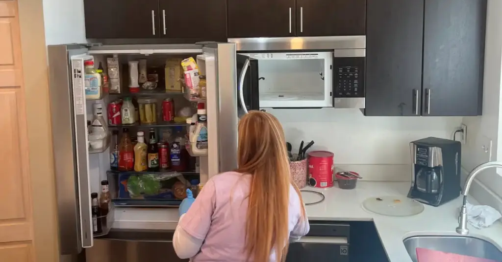 Person with long hair stands in front of an open, full refrigerator in a modern kitchen with dark cabinets.