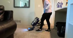 Person in a pink shirt and blue gloves pushes a floor-cleaning machine across a beige carpet in a living room.