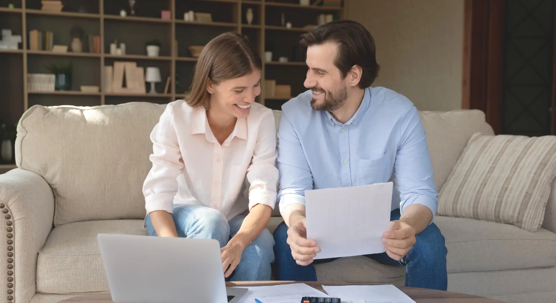 Smiling couple sits on a couch with a laptop and papers, appearing to review documents together.