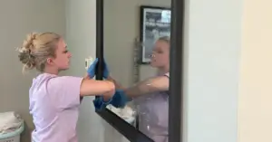 Woman cleaning a large mirror with a blue cloth, her reflection visible in the glass.