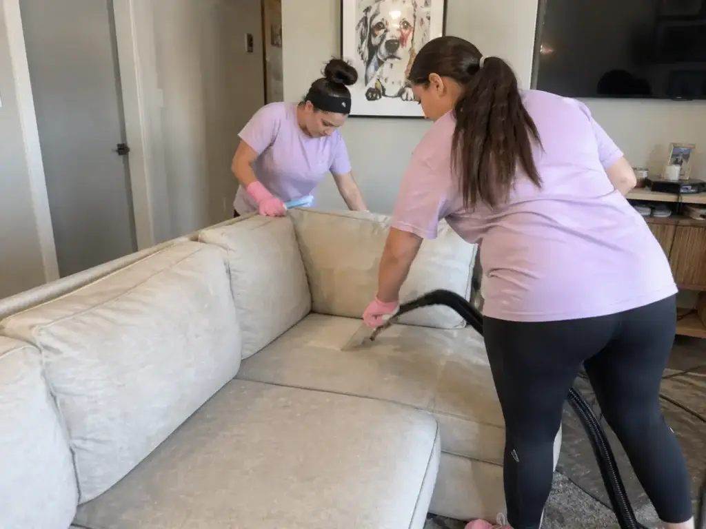 Two women in purple shirts cleaning a beige couch with a vacuum in a living room.