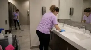 Two people in purple shirts clean a public restroom; one wipes the counter, the other mops the floor.