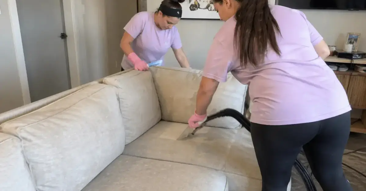 Two women in purple shirts clean a beige couch; one vacuums while the other adjusts a cushion. - Jeannie’s Cleaning Company Two women in purple shirts clean a beige couch; one vacuums while the other adjusts a cushion.