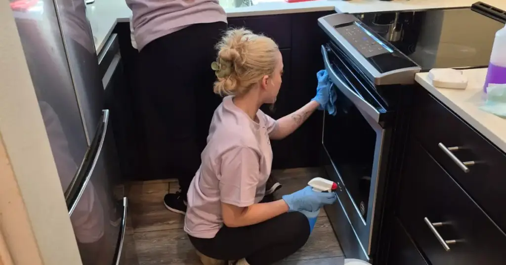 Two people in gloves cleaning a kitchen, with one wiping an oven door and the other cleaning the counter.
