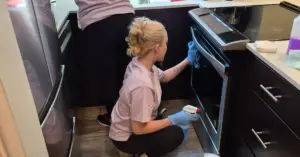 Two people in gloves cleaning a kitchen, with one wiping an oven door and the other cleaning the counter.