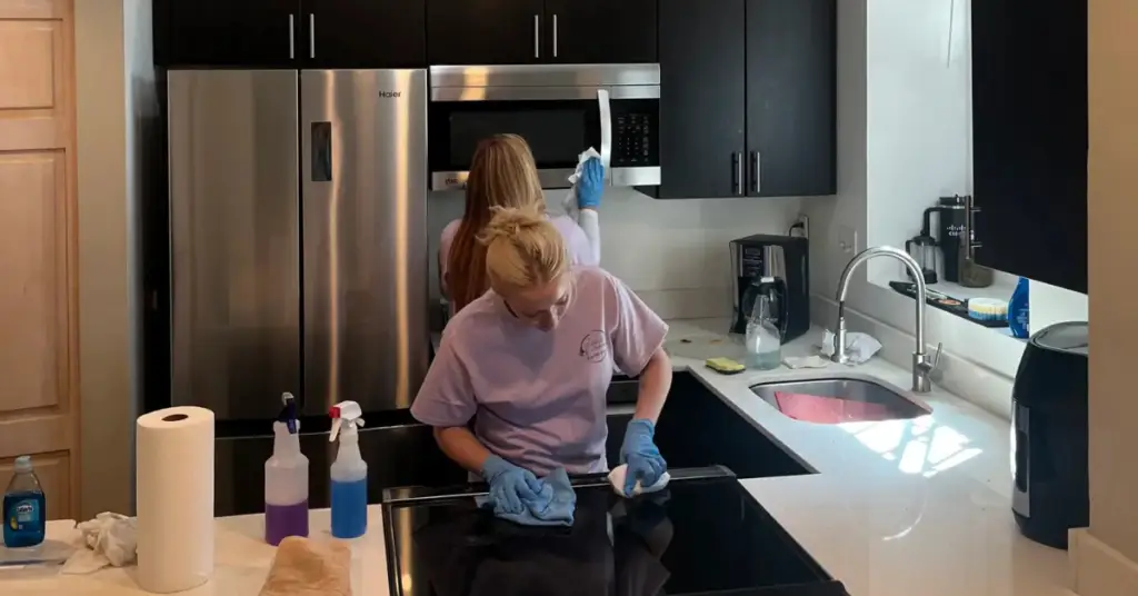 Two people wearing gloves clean a modern kitchen, wiping the stovetop and a microwave above the stove.