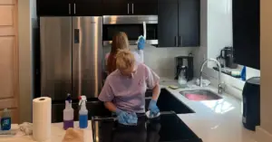Two people wearing gloves clean a modern kitchen, wiping the stovetop and a microwave above the stove.