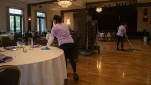 Two cleaners in purple shirts reset a banquet hall after an event, wiping tables and mopping the floor